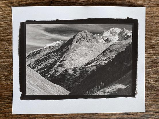 Phone picture of a 8x10" piece of paper with a slightly smaller kallitype print of a high alpine mountain landscape. The picture shows the view of deep valley with a long smooth glacial ridge leading up to snow covered peaks. A band of forest is running down the slopes in the foreground, still mostly in the shade, but a small area near the edge which is catching the morning sun. 