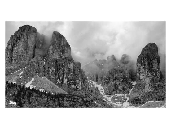 Wide-format B&W photo of a classic Dolomites landscape with steep couloir (still with some snow) going up between massive rock towers & needles, their tops slighly obscured by a layer of thick moody gray clouds.