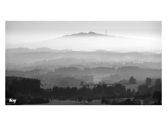 B&W wide-format photo of a hilly landscape in layers of fog and a larger mountain rising up behind a major fog layer in the distance. Only its built-up summit part (with churches, TV tower and other buildings) is visible. Layers of forest and fields in the lower foothills in the foreground.