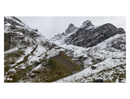 Wide-format photo of a high alpine landscape dusted in a thin layer of snow, amplifying the rich textures and last remaining bits of color. A large and deep erosion chute is curving up steeply towards the ridge line and large rock peaks higher up. Large vertical rock faces on the right hand side. Gray sky completely filled with snow clouds.