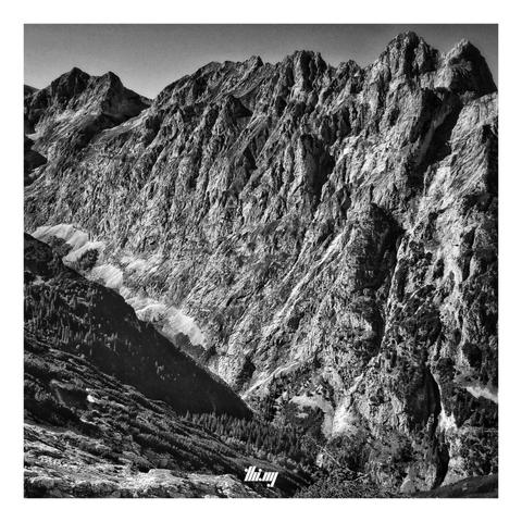 Square format high contrast B&W photo of a large and very deep high alpine valley, looking down from a mountain pass around 800 meters above the valley floor. A small footpath with switchbacks through some forest is visible near the bottom. The opposite side of the valley is formed by a single massive rock face with jagged peaks & ridge line...