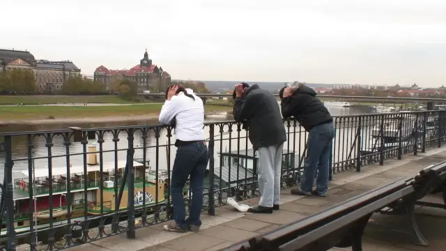 Three people leaning on a metal railing near the river Elbe in the center of Dresden. Each person is using their hands to cover their ears, elbows on the metal. Some large historical buildings in the background

Image credit: Markus Krison