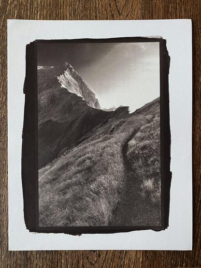 Phone picture of a 8x10" sheet of paper with a smaller kallitype print of a narrow path leading to a high alpine mountain pass in front of a sharp ridge leading up to a large mountain peak. Intense bright light from the other side of the pass is creating a high contrast.