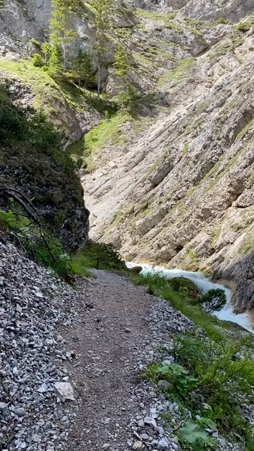 First person video of someone walking on a narrow path near a fast flowing mountain creek inside a deep canyon surrounded by steep cliffs. The water color is an intense milky light blue/cyan...