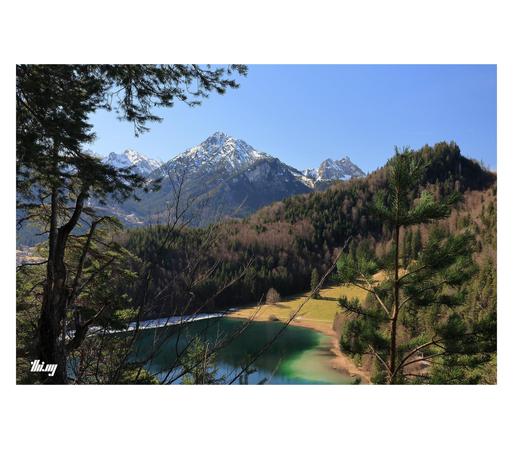 View of a mountain landscape from above a small lake with green/blue water, surrounded by forest and meadows. Snow capped larger mountains in the background.