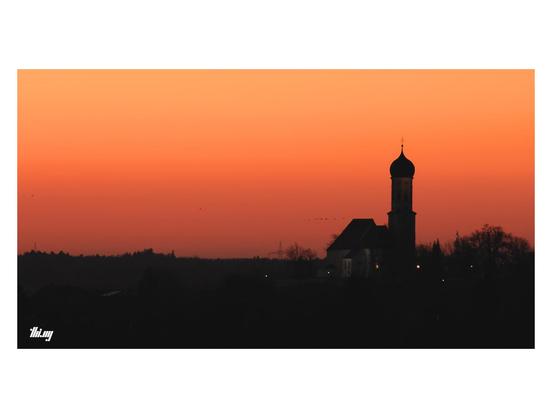 The silhouette of a typical Bavarian church with onion dome and the surrounding tree line against a deep orange/red sky. Cloudless sky.