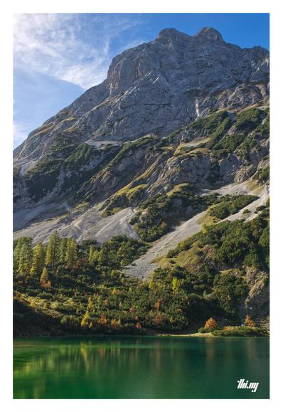 A mountain lake with deep green water in front of a steep talus slope and vertical rock face going up hundreds of meters directly behind. The lower parts near the lake are covered in dense bushes of dwarf mountain pines and some larches, all faintly reflected on the lake surface. The beautiful sunny day brings out the autumn colors with some of the larches in bright orange. Semi-overcast blue sky.