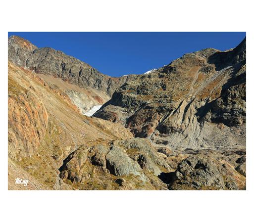View of a high alpine section of a glacial valley approx. 1 km from the current glacier tongue (a fragment of it is visible in the background). Rocks of many different colors and traces of glacial activity and heavy erosion are forming an chaotic looking landscape. Harsh shadows are emphasizing the various features. The plain blue sky gives this alien looking landscape an extra abstract element.