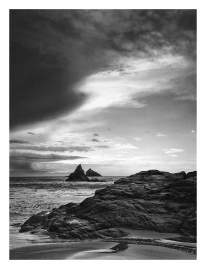 B&W photo of a large wet slab of rock in the sand near the waterline and the silhouettes of some sea stacks/needles further out. Half of the sky is covered by a moving storm front moving south, creating an eerie light.
