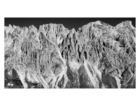 Wide-format B&W photo of a long vertical craggy rock face with extreme folds in the rock. Below steep talus fields, partially overgrown with grass and some small patches of dwarf mountain pines.