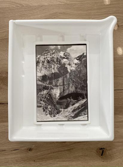 A ~10x7 inch kallitype print of a mountain landscape (portrait aspect ratio) in a slightly larger white photo processing tray on a wooden floor.