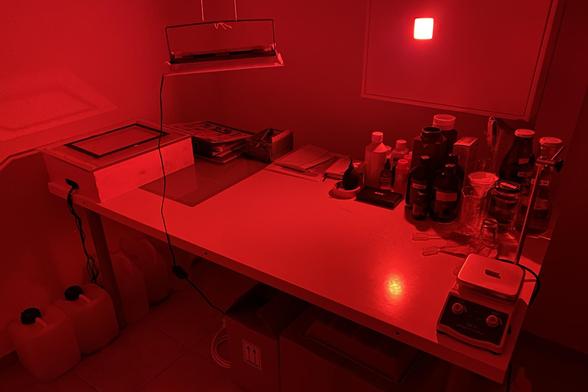 Photo of a table with various bottles, beakers, heating plate mixer, papers and other small items. All illuminated only by a small red LED light.