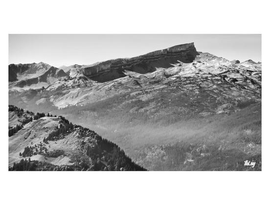 Wide-format B&W photo of a sloped mountain massif in the distance and a large rocky plateau in front, transitioning into forests further down the valley. The edge of a closed mountain ridge in the bottom-left corner.