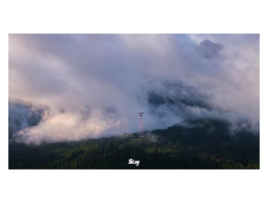 Photo of a dramatic cloud forming from the dense rising fog near the steep slopes of the Zugspitze (its summit visible in a gap between the clouds). The cloud is illuminated by pink/orange pastel early morning light. A massive single red-white mast for a cable car is towering above the forest in the foreground, some nearby trees also catching the light of the rising sun. In the background, glimpses of talus fields through the clouds.