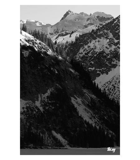 B&W photo of a frozen mountain lake surrounded by steep, almost vertical rock faces and snowy mountain peaks. Only the tops are catching the last light, the rest in deep shade...