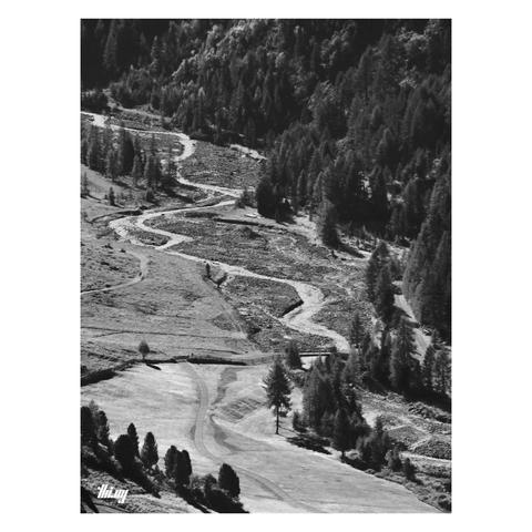 B&W photo of a mountain creek meandering through a much larger rocky river bed in a valley. One side of the valley is covered in dense forest. A path is winding its way through meadows near the creek on the other side.