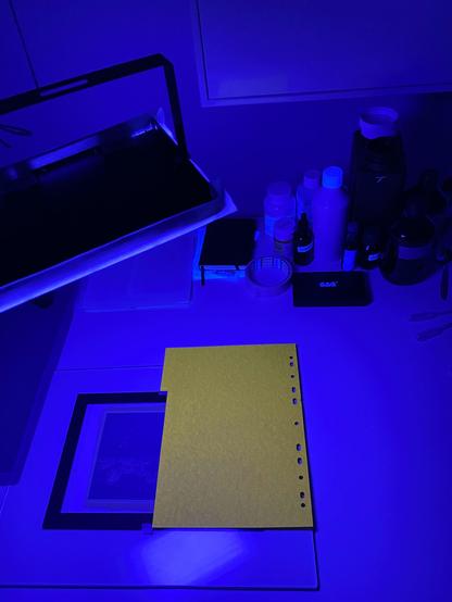 A white table under a strong UV light which is used to expose a kallitype print underneath a thick piece of glass, half covered by a yellow piece of card. Various bottles of photo chemicals are standing in the corner...