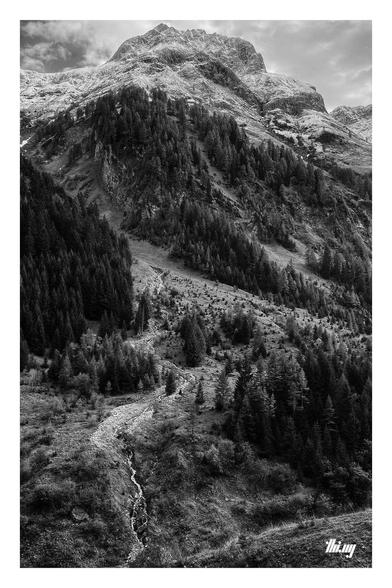 B&W photo of a view across a beautiful alpine valley and the semi-forested slopes and rocky peaks on the other side of the valley, the higher up parts slightly sprinkled with fresh snow. A small creek and waterfall coming down through one of the gullies. Cloudy, wintry sky.