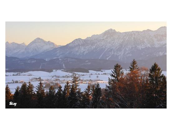 Wide-format view of a small section of the Northern Alps and their foothills during sunset on a bright winter day. The sky is glowing in a mix of pastel yellows, orange, cyan. Snowy mountains in the back and white fields closer in the foreground. Closest to the camera are the tops of a patch of forest, the trees illuminated in warm red light. A sun beam is shining on a small village in mid distance.