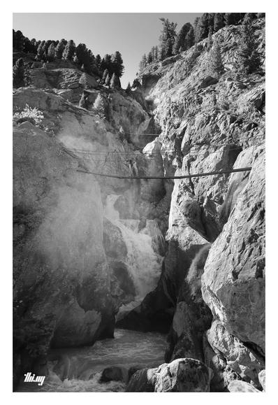 B&W photo of a multi-stage waterfall crashing down a deep and narrow rocky canyon, intense spray developing in the lower parts. A hiking/climbing path is zigzagging a few hundred meters up across the canyon via precarious looking small rope bridges. At the top edge/horizon, small patches of forest.