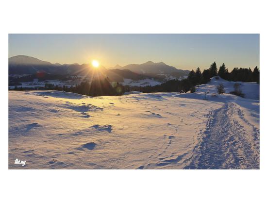 Sunset over a beautiful snowed in landscape in the foothills of the Alps on a cold and crisp clear day. The sun flares just before disappearing behind the ridges of the mountains in the distance, which are already casting their shadows far and long. The view is from an open ridge and the snow in the foreground is illuminated by orange light. Other people's foot steps have a created a path in the snow, leading into the distance towards a forest on the right hand side. Clear azure sky.