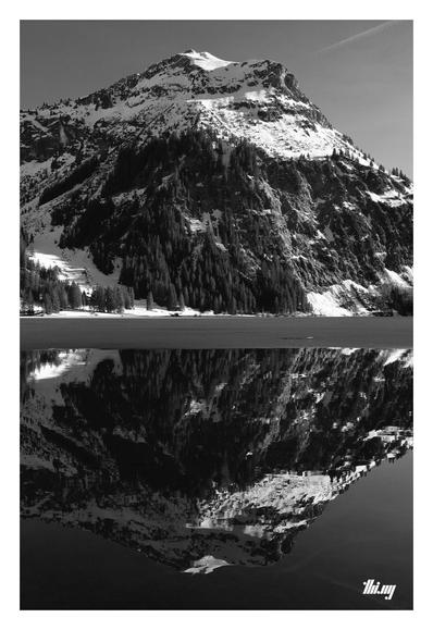 B&W photo of a large snow covered mountain reflecting on the completely still (and partially ice covered) surface of the lake in front of it. 