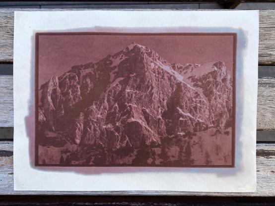 Photo of a failed Kallitype print of a large mountain peak. Instead of black and white, the print is strong burgundy, with slightly blueish highlights. The emulsion outside the frame is continuing to bleed further into the surrounding paper