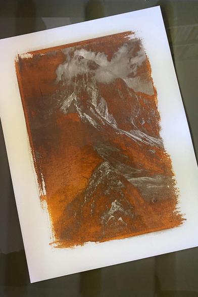 A kallitype print of a mountain peak during early stages of development, still immersed in developer in a photo tray. The darkest parts of the image have become solarized and turned into shades of saturated rusty orange, whilst the lighter parts of the image can still be made out (ridges and clouds)
