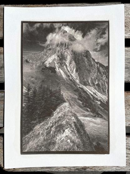 Phone picture of a 8x6" kallitype print of a large mountain peak partially shrouded in clouds forming around it. In the foreground is a narrow ridge with a very similar profile as the peak. A small group of low-growth trees next to it and a large valley running down and opening up from the pass on the left. Dark moody sky.