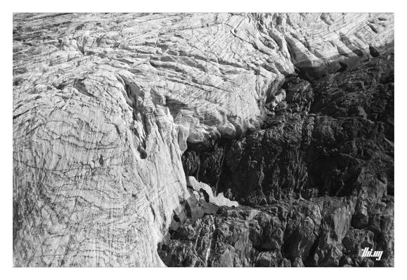B&W close up of a glacier edge around an island of dark exposed bedrock, the melt water forming small waterfalls. The glacier ice is still dozens of meters thick with small ceracs and dark contour lines. The more rock is exposed, the more heat is accumulated in summer and the faster the glacier is shrinking... a vicious circle!