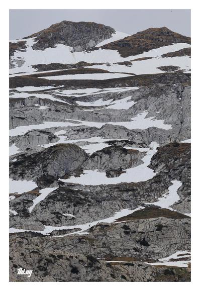 Abstract photo of a postglacial landscape with layers of gray rocks and interspersed snowfields, some patches of old brown grass. Plain gray sky.
