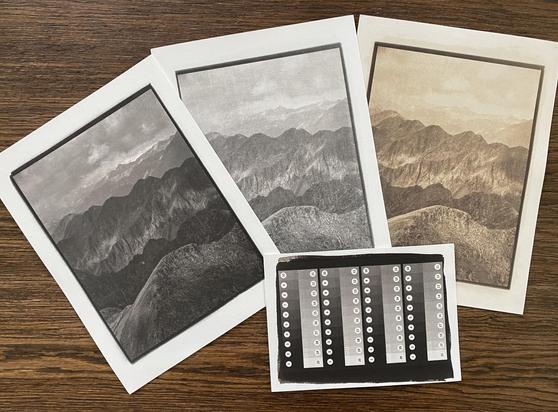 Three kallitype prints of a barren mountain landscape lying on a table. Each print has a different tone (grays and sepia) and contrast. A smaller kallitype print is next to them, showing swatches to verify tone curves at four different exposure times.