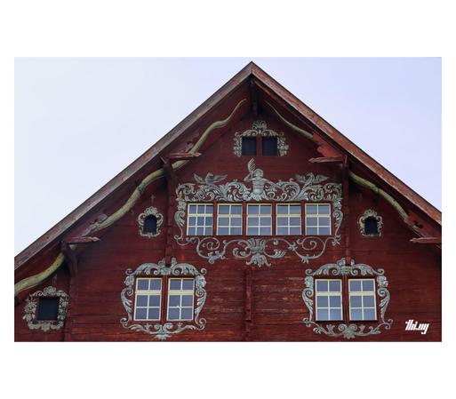 View of the wooden gable of a 16th century rustic house. The planks have been painted in reddish brown and the areas around the windows decorated with gray ornaments. The painted tails of two large snakes are running down along the inside of the overhanging roof. Clear azure sky.
