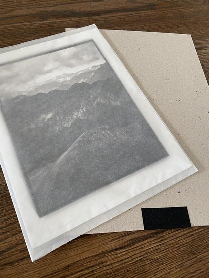 A 8x6" kallitype print inside a parchment pouch lying atop of two sheets of thick speckled gray cardboard with a hinge made of black textile tape