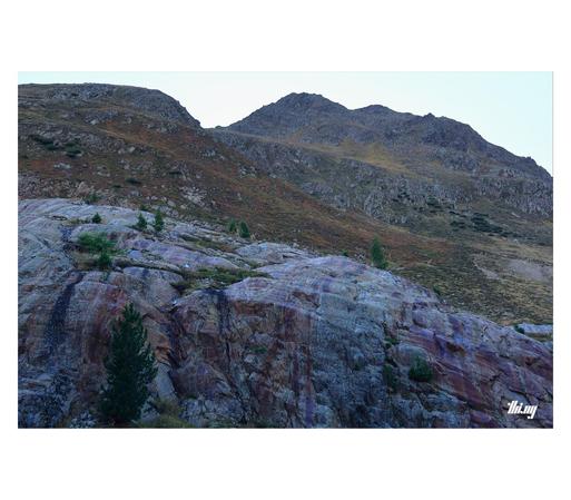 View of a high alpine post-glacial landscape with the bottom half entirely consisting of an exposed, colorful section of bedrock (grays, purples, dark reds), sharply delineated from the surrounding landscape, smoothed by abrasion of the former glacier here and indicating its previous vertical extent. Above that large bare rock face are alpine meadows and tundra vegetation in beautiful autumn colors. Clear bright sky.