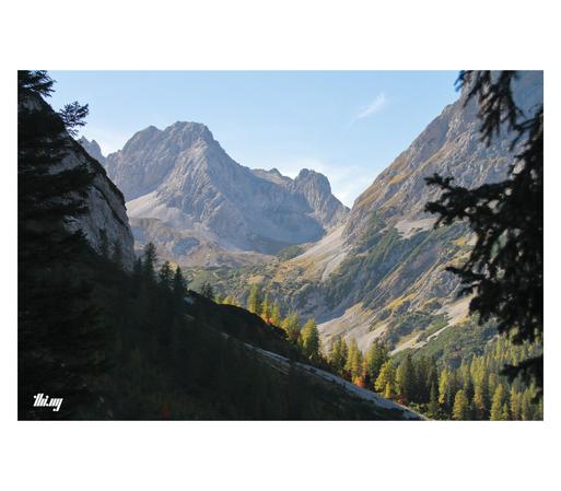 View of a beautiful high alpine valley with the lower parts covered in larch forest, the trees in beautiful autumn colors, higher up patches of dried grass and mountain dwarf pines among talus fields and steep large rocky peaks. Clear azure hazy sky. The silhouettes of some tree branches in the foreground are framing the scene left/right.