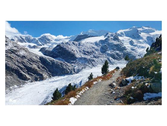 A short section of path visible a few hundred meters above a large, but dying glacier. Snow and ice packed 4000 meter mountain peaks in the back, other glaciers coming down in side valleys, but disconnected (too short) due to climate change. The path is surrounded by tundra vegetation in beautiful autumn colors. Blue sky with some cumulus forming on the horizon.