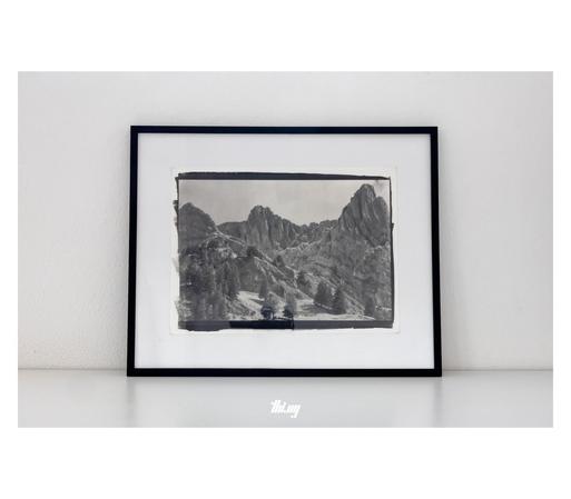 A 15x11" framed kallitype print leaning against a white wall on a white semi-glossy surface. The print itself is of a high alpine landscape with extreme rock folds groups of trees.