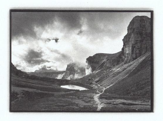 Scan of a 7x5" kallitype print of a moody alpine landscape. A small hiking path leading into the background at the feet of talus fields and past a small mountain lake. Big dark clouds moving through the layers of rock towers