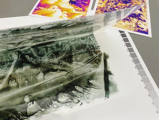 Photo of a folded and then back half-opened sheet of transparency film with black ink traces forming an abstract texture exhibiting intense viscous fingering. In the back two color prints (in a "thermal camera" style color theme) showing simulated variations of the same overall effect.