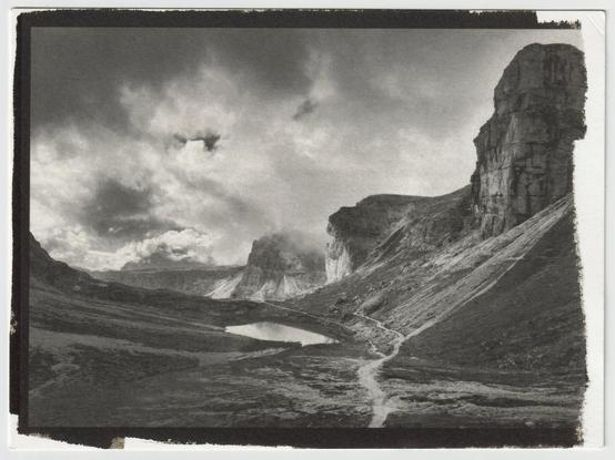 Scan of a 7x5" kallitype print of a moody alpine landscape. A small hiking path leading into the background at the feet of talus fields and past a small mountain lake. Big dark clouds moving through the layers of rock towers
