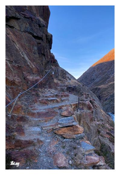 POV view of a narrow path with natural rock steps along the side walls of a canyon (near its entrance). The section is partially secured with a steel rope along the rock face on the left. Clear blue sky with the opposite side of the valley catching the last sun light of the day.