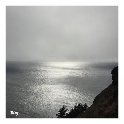 Square photo of a calm, almost empty ocean landscape with the silhouette of a steep slope of the Cape Lookout peninsula in the bottom-right corner. The top part of the image is an almost uniform gray, the sun slowly trying to break through the thick fog, causing a silvery light spectacle on water below.