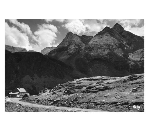 B&W photo of a grand high alpine mountain landscape. A small road leading through grassy terrain in the foreground to a hut in a bend, after which the road is disappearing from view. Dark steep slopes with talus fields leading up to the large massif behind. Large & bare pyramidal rock faces higher up and layers of other peaks in the back. Semi-cloudy sky.