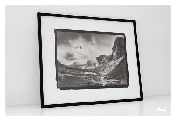 Photo of the same print, this time framed in a 50x40cm thin black frame, leaning against a white wall.