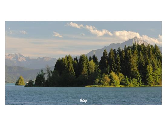 Wide-format photo of a large lake in the pre-Alps. A group of trees on a small peninsula, illuminated by the warm evening sun, further back a small island. In the background the mountains of Tyrol, some still with patches of snow. Hazy sky with some clouds.