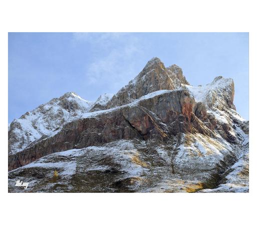 Photo of beautiful multi-layered mountain peak dusted in snow, some of its slopes catching the morning light. The a band of rusty red rock is running across the view just above the more gentle slopes below.