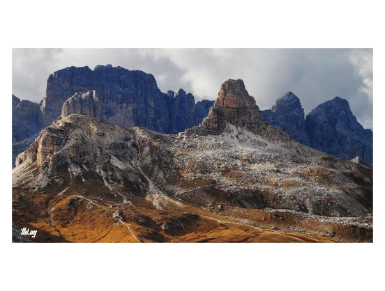 View of a narrow rocky ridge with a large rock tower in front of another mountain range in the background. Small paths are leading through talus fields towards the base of the tower. The entire scene is bathed in the warm late afternoon light of October. The grass patches have turned into a rich orange brown. Cloudy sky.