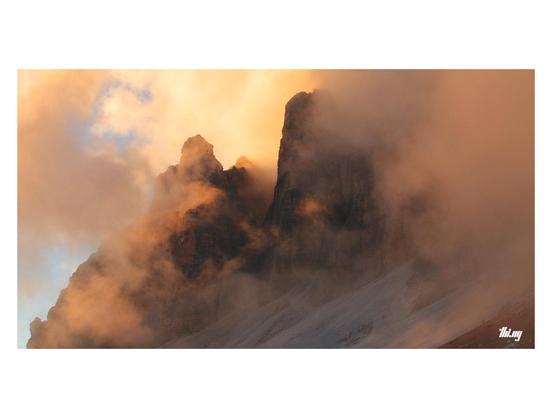 Wide-format view of dense clouds forming around some vertical rock towers during sunset, the red orange light refracting in the mist and making the sky glow.