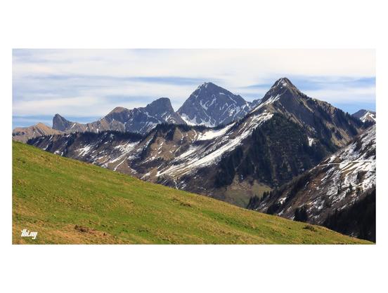 Wide-format photo of an alpine landscape in early spring. Unlike the other mountain slopes, the meadow in the foreground is already snow free and showing signs of first green grass growing. Further in the back, layers of both smooth and craggy ridges, still semi-covered in patches of snow. Overcast hazy sky.